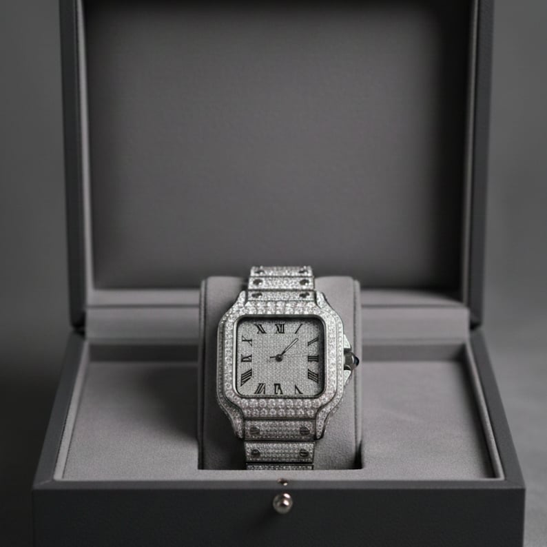 May include: A silver wristwatch with a square face and a matching band, encrusted with small, sparkling stones. The watch is displayed inside a gray box with a hinged lid, set against a gray background. Roman numerals mark the hours on the watch face.