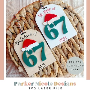 Könnte beinhalten: Zwei Weihnachtsschmuckstücke mit dem Text "The Season of 67 2025" und "The Year of 67 2025". Jedes Schmuckstück hat einen roten Weihnachtsmannhut und grüne Zahlen. Die Ornamente befinden sich auf einem geflochtenen Korb.