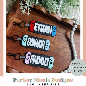 May include: Three black keychains with names: Ethan, Connor, and Makinley. Each keychain has a black base with a colourful game controller design and the name in white. The keychains are on a wooden tray.