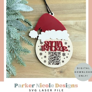 以下が含まれることがあります： サンタの帽子と円形のベースの形をした木製のクリスマスオーナメント。オーナメントには、赤い「SANTA TRACKER」の文字、雪の結晶、QRコードが描かれています。帽子は赤色で、白いトリムが付いています。吊り下げ用の黒い紐が付いています。