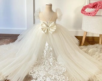 May include: A cream-colored floor-length dress featuring a voluminous tulle skirt, ruffled cap sleeves, and a sheer bodice. The back is adorned with a large satin bow at the waist and an intricate white floral lace applique trailing down the center.