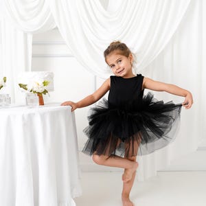 Może przedstawiać: Młoda dziewczyna w czarnej sukience tutu. Sukienka bez rękawów ma dopasowany gorset i pełną, warstwową spódnicę z tiulu. Jest boso i stoi na jednej nodze, uśmiechając się. Biały obrus i zasłony są w tle.