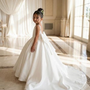 May include: A floor-length white sleeveless dress featuring a wide scoop neckline, a fitted bodice, and a full, voluminous skirt with a long train. A large, structured white bow is attached at the center back of the waistline.