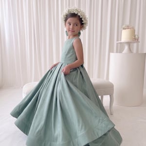 May include: A floor-length sage green sleeveless dress with a fitted bodice and a full, pleated skirt. The dress features a wide hemline and a clean, structured silhouette. A delicate white floral crown is worn on the head, adding a soft contrast.