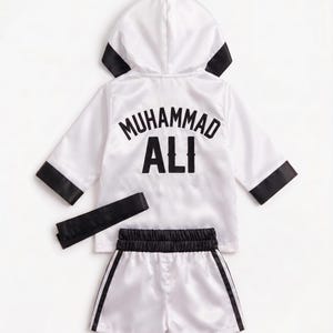 Könnte beinhalten: Ein weißer Boxmantel aus Satin mit Kapuze, schwarzen Ärmelabschlüssen und einem schwarzen Gürtel. Auf dem Rücken befindet sich der Schriftzug MUHAMMAD ALI in schwarzen Buchstaben. Dazu gehört eine passende weiße Boxershorts aus Satin mit schwarzen Seitenstreifen.