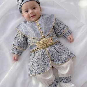 May include: A baby wearing a traditional outfit with a patterned jacket, hat, and belt. The jacket is blue and white with gold trim and buttons. The outfit includes white pants and shoes. The baby is smiling.
