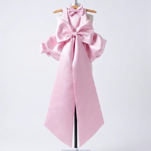Puede incluir: Un vestido sin mangas de satén rosa claro que presenta un corpiño estructurado con una pajarita a juego en el escote. Un lazo grande y prominente está centrado en la cintura, con dos paneles de tela largos y anchos que bajan hasta el dobladillo.
