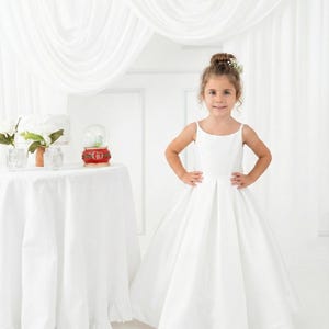 May include: A floor-length white dress with thin spaghetti straps and a pleated skirt. The dress features a simple bodice with a straight neckline and a full, flowing skirt that reaches the floor, creating a clean and structured silhouette.