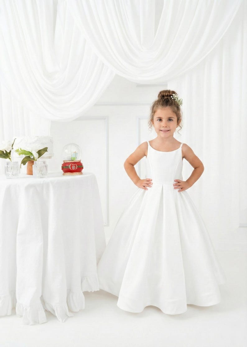 May include: A floor-length white dress with thin spaghetti straps and a pleated skirt. The dress features a simple bodice with a straight neckline and a full, flowing skirt that reaches the floor, creating a clean and structured silhouette.