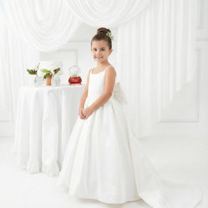 May include: A floor-length white dress with thin spaghetti straps, a fitted bodice, and a full skirt featuring a large bow at the back. The dress has a long train that extends across the floor, creating a formal silhouette with clean lines.