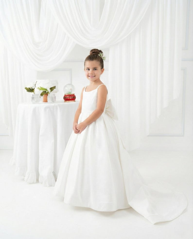 May include: A floor-length white dress with thin spaghetti straps, a fitted bodice, and a full skirt featuring a large bow at the back. The dress has a long train that extends across the floor, creating a formal silhouette with clean lines.