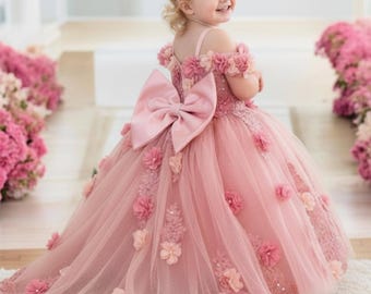 May include: A dusty pink floor-length dress featuring off-the-shoulder sleeves, a large satin bow at the back, and a tulle skirt adorned with three-dimensional fabric flowers and lace appliqués. A matching floral headband completes the ensemble.