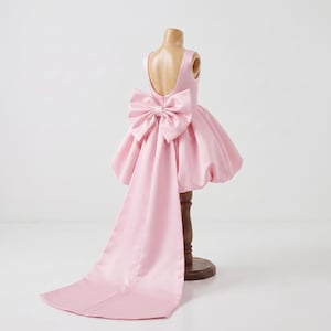 Puede incluir: Un vestido rosa claro sin mangas con escote redondo en la espalda y una falda abullonada voluminosa. Un lazo de satén grande y estructurado está sujeto a la cintura, con una larga cola fluida que se extiende desde el lazo hasta el suelo.