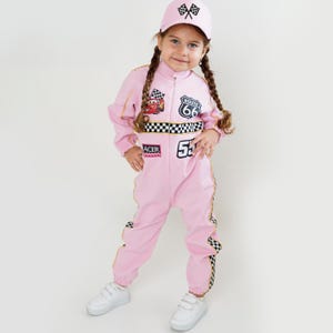 Puede incluir: Mono de carreras rosa con detalles a cuadros blancos y negros y el número 53. El atuendo incluye una gorra a juego con banderas a cuadros. El mono presenta un parche de la Ruta 66 y un gráfico de coche de dibujos animados.
