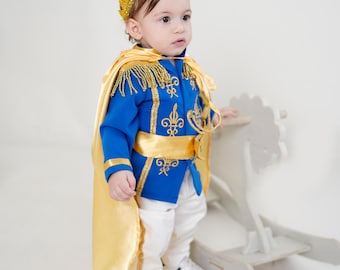 May include: A royal-inspired costume featuring a bright blue jacket with gold embroidery and fringe epaulettes, a matching gold satin cape, a gold satin sash, white trousers, and a gold crown. The outfit is worn with white trainers.
