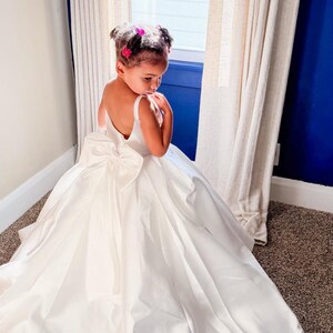 May include: A floor-length white dress with a sleeveless bodice, a deep scoop back, and a large, prominent bow centered at the waistline. The skirt is voluminous and full, featuring a smooth, satin-like fabric that drapes to the floor.