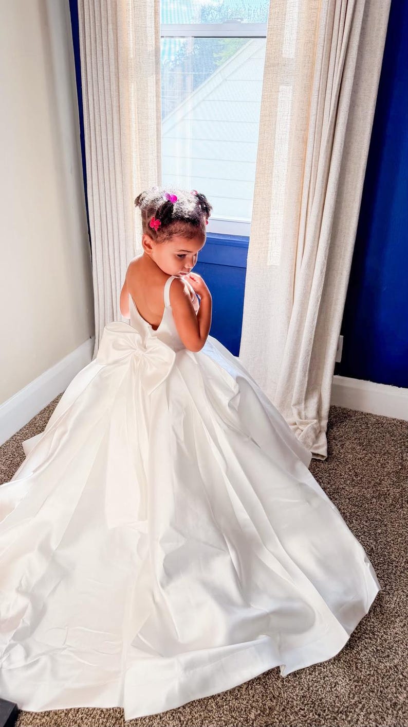 May include: A floor-length white dress with a sleeveless bodice, a deep scoop back, and a large, prominent bow centered at the waistline. The skirt is voluminous and full, featuring a smooth, satin-like fabric that drapes to the floor.