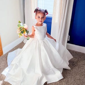 May include: A floor-length white sleeveless dress with a fitted bodice and a full, pleated skirt. The dress features a wide, rounded neckline and a smooth, satin-like finish. A small bouquet of artificial flowers with pink and cream petals is held.