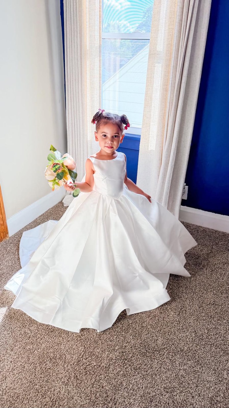May include: A floor-length white sleeveless dress with a fitted bodice and a full, pleated skirt. The dress features a wide, rounded neckline and a smooth, satin-like finish. A small bouquet of artificial flowers with pink and cream petals is held.