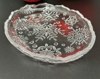 May include: Clear glass serving dish with snowflake design. The oval-shaped dish has a scalloped edge and is decorated with etched snowflakes. Two red Christmas ornaments are in the background.