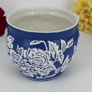 May include: A blue ceramic planter with a white floral design. The planter has a wide, open top and a smooth, glossy finish. The white floral pattern is raised and adds texture to the planter's surface. The interior is white.