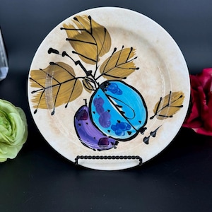 May include: A round, off-white ceramic plate with a hand-painted design of two plums and leaves. One plum is blue, the other purple, with brown and gold leaves. The plate is displayed on a black metal stand.