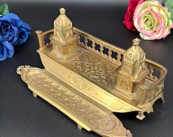 May include: A brass inkwell set featuring two domed towers and a rectangular base with intricate geometric engravings. The set includes a separate, elongated tray with matching ornate patterns, all crafted from a metallic gold-toned material.