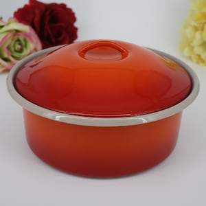Może przedstawiać: Okrągły, pomarańczowy garnek emaliowany z dopasowaną pokrywką i srebrną obwódką. Pokrywka ma mały uchwyt. Garnek stoi na białym tle z czerwonymi i żółtymi kwiatami.