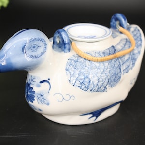 May include: A ceramic duck-shaped teapot with blue and white floral patterns. The teapot has a spout, a small opening on top, and a woven handle. The duck's head and wings are detailed with blue paint.
