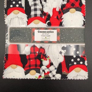 May include: A stack of square fabric pieces featuring a gnome pattern with red, black, and white colors. The fabric is wrapped in a paper band labeled Gnome-antics, 5 Karat Crystals, and Wilmington Prints. The edges of the fabric squares are pinked.