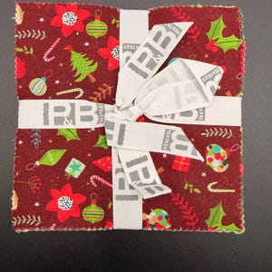 May include: A square stack of fabric squares tied with a white ribbon featuring the text P&B TEXTILES. The fabric has a dark red background with a repeating Christmas pattern of green pine trees, red poinsettias, holly leaves, and ornaments.