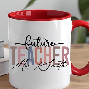 May include: White ceramic mug with a red rim and handle. The mug has the text "future teacher" in black script with the word "teacher" in colorful letters. The text "Miss Smith" is written in black script below the word "teacher".