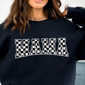 May include: Black sweatshirt with the word "MAMA" in large, white, checkered letters across the chest. The sweatshirt has a crew neck and long sleeves. The image is a close-up of the sweatshirt.