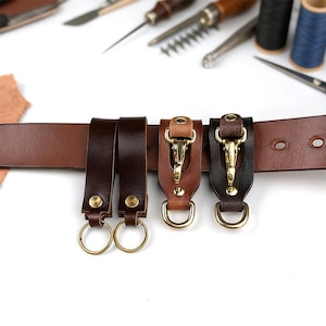 May include: A brown leather belt with three key holder attachments in varying shades of brown and black. Each attachment features a brass clasp and a D-ring. Two key holders have gold-colored rings. The background includes leather tools.