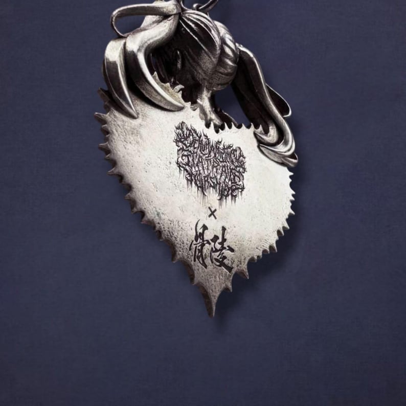 May include: A silver-toned pendant with a heart-shaped saw blade design. The pendant features a stylized design and text in the center and at the bottom. The pendant is suspended from a loop at the top.