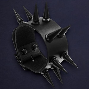 May include: A black leather wristband featuring several black conical spikes. The wristband has a buckle fastening and is displayed against a dark blue backdrop. The spikes are uniformly spaced, giving a bold, edgy appearance.