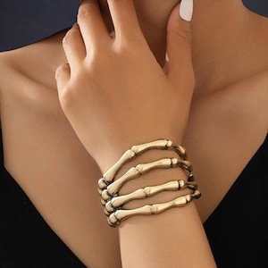 May include: A gold-colored bracelet shaped like a skeleton hand. The bracelet is worn on a wrist and is made of a metallic material. The bracelet has a detailed design, with each finger bone clearly defined. The overall style is gothic and edgy.