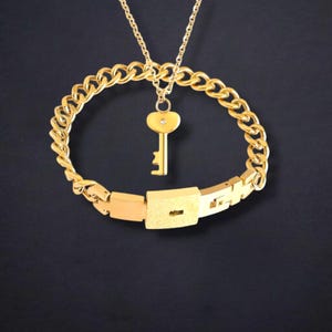 May include: A gold-coloured necklace with a key pendant and a matching bracelet. The key pendant has a heart-shaped top with a small gem. The bracelet features a lock design. The jewellery is set against a dark background.