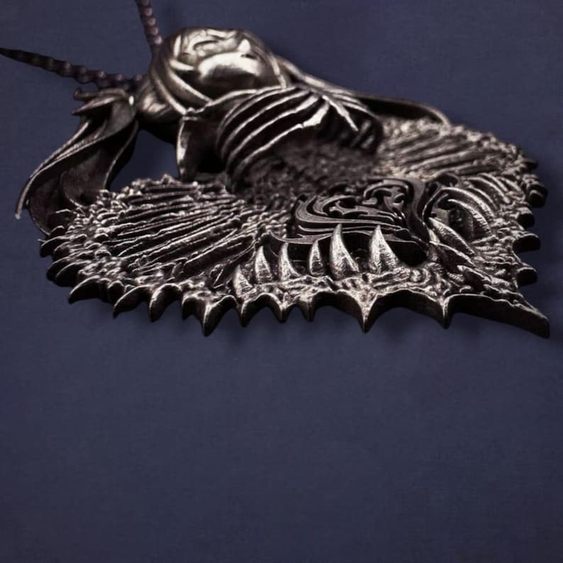 May include: A detailed, silver-toned pendant featuring a mythical creature design. The pendant showcases a textured surface with intricate details, including a horned figure and a spiky, wing-like structure. The pendant is suspended from a chain.