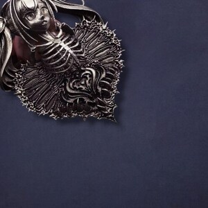 May include: A detailed, silver-toned pendant featuring a stylized female figure with a skeletal ribcage design. The pendant has intricate, ornate detailing and is set against a dark blue background. The design has a gothic or fantasy theme.