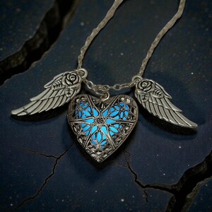 Pu&ograve; includere: Una collana con ciondolo a forma di cuore argentato con un centro blu brillante e due ciondoli a forma di ali. Il cuore ha un design a filigrana. Le ali e la catena sono anch'esse argentate. La collana &egrave; esposta su una superficie blu scuro.