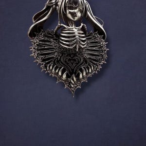 May include: A silver-toned pendant featuring a stylized figure with long hair and a skeletal torso. The figure is surrounded by a heart-shaped, ornate design with sharp edges. The pendant is set against a dark blue background.