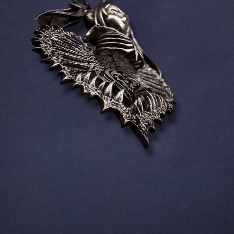 May include: A detailed, silver-toned pendant with a gothic design. The pendant features a central figure with a hood, surrounded by intricate, spiky, and textured elements. The background is a solid, dark blue.