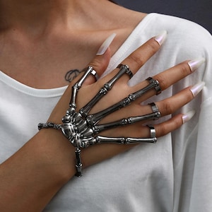May include: A silver-toned skeleton hand bracelet with rings on each finger. The bracelet is attached to the hand with a series of articulated bones, creating a gothic and edgy aesthetic. The rings are simple bands, complementing the skeletal design.