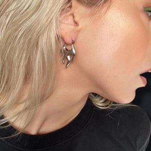 May include: A silver-colored earring with a flame design is shown. The earring is a hoop style with a fluid, flame-like shape. The earring is worn on the ear of a person with blonde hair and a black shirt.