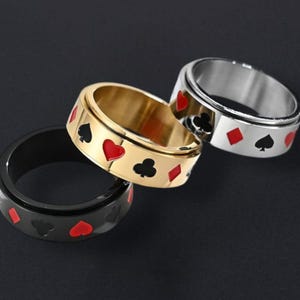 May include: Three rings featuring playing card suit symbols. One ring is black with red symbols, another is gold with red and black symbols, and the third is silver with red and black symbols. The rings are displayed on a dark background.