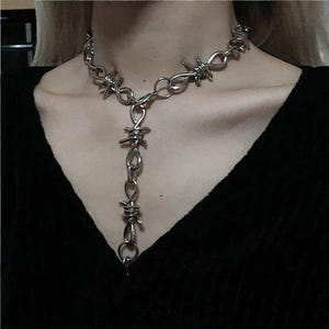 May include: A silver-colored necklace with a barbed wire design. The necklace has a choker-style chain with a vertical drop featuring more barbed wire elements. The jewelry is worn against a black top.