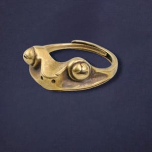 May include: A brass-coloured ring with an adjustable band. The ring has a stylized design with rounded elements and a textured surface. The design includes two raised circular features and a central, curved shape. The ring is set against a dark blue background.