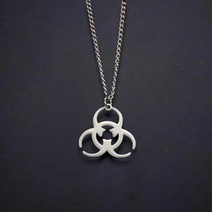 May include: A silver-colored pendant necklace featuring a biohazard symbol. The pendant is suspended from a delicate chain. The necklace is displayed against a dark background.