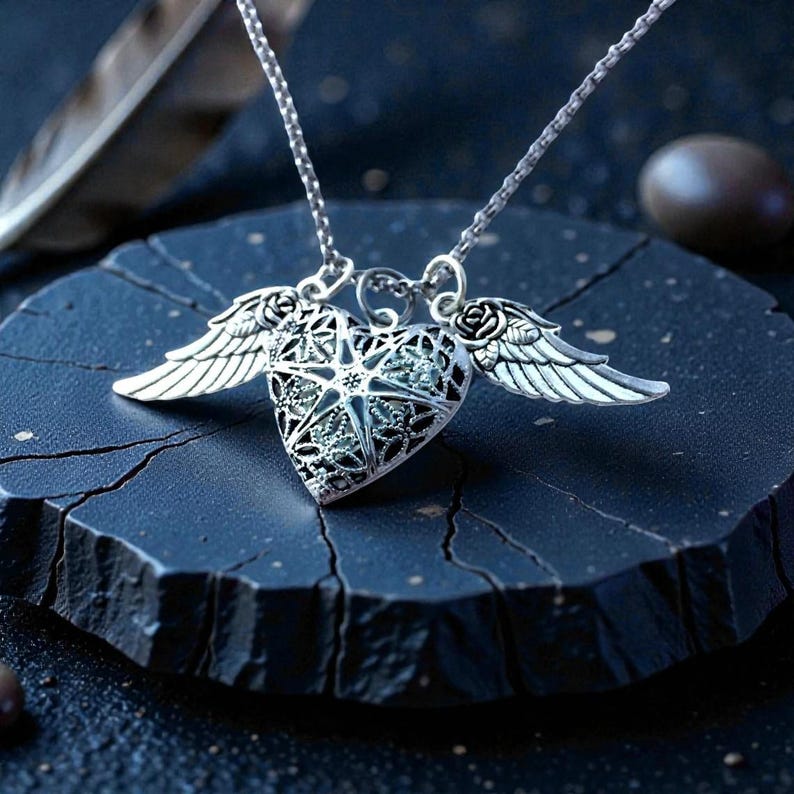 Pu&ograve; includere: Collana con pendente a forma di cuore argentato con un intricato design traforato, ornato da piccole ali su entrambi i lati. Il pendente presenta un motivo a stella ed &egrave; sospeso a una delicata catena. La collana &egrave; esposta su una superficie blu scuro strutturata.
